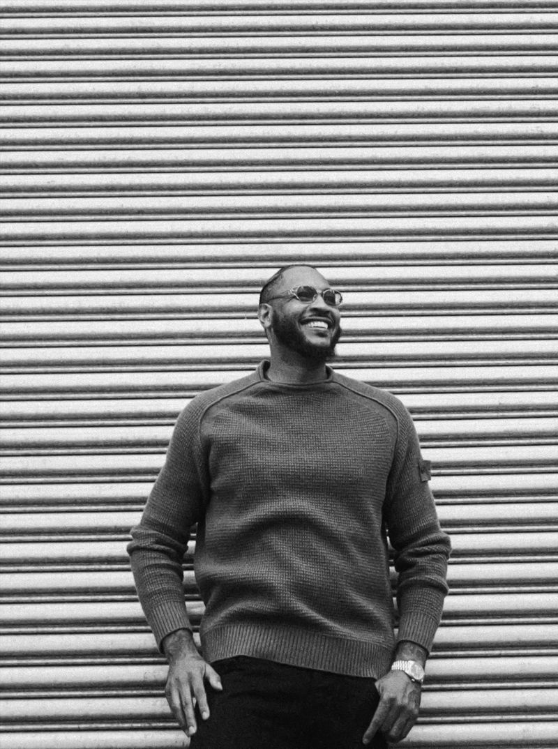 Black and white medium shot of a bearded man in glasses and a sweater smiling, posing in front of a corrugated metal wall.