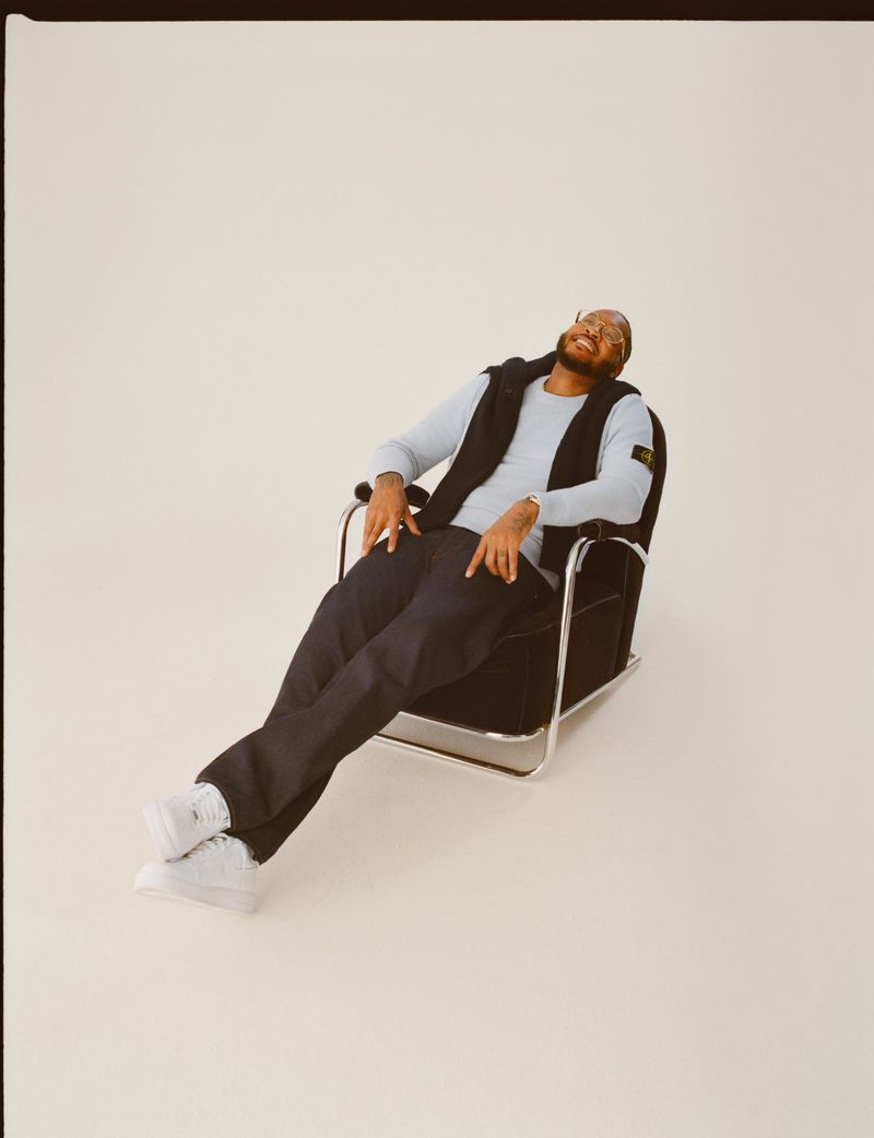 Man laughing while seated in a black armchair against a pale studio backdrop wearing a light sweater and dark trousers