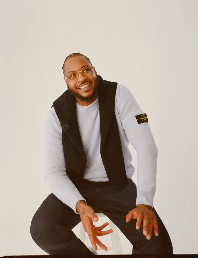 Portrait of a smiling adult man in a light-blue sweater and black vest, seated against a neutral studio backdrop, looking up and to the left.