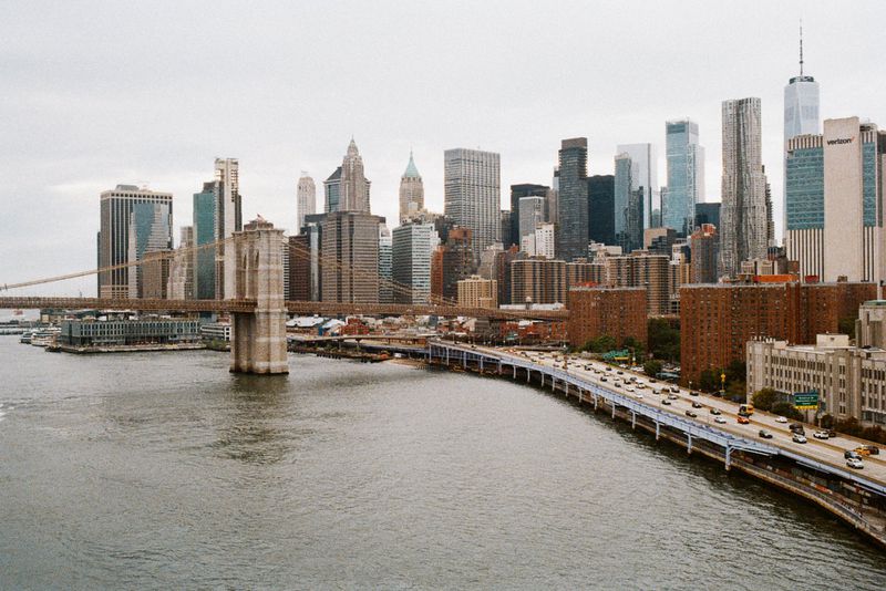 Daytime cityscape featuring the Brooklyn Bridge connecting to Manhattan, New York, with numerous skyscrapers and river views.