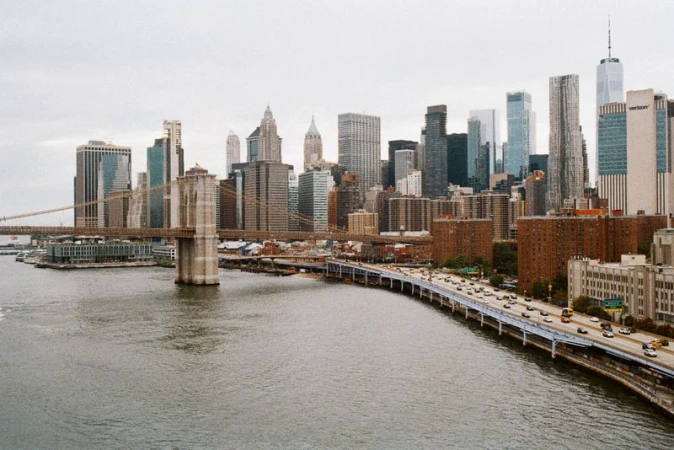 Daytime cityscape featuring the Brooklyn Bridge connecting to Manhattan, New York, with numerous skyscrapers and river views.