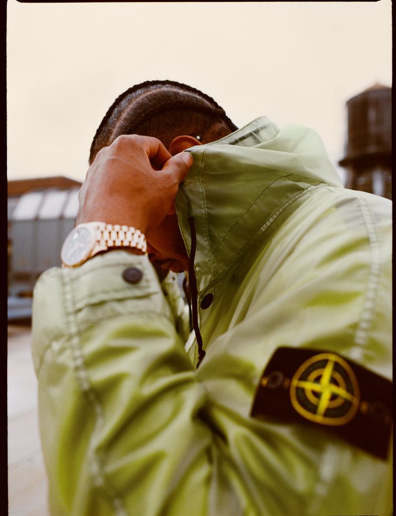 Close-up shot of person wearing a light green hooded jacket with a Stone Island logo patch, gold watch, and an urban setting background.