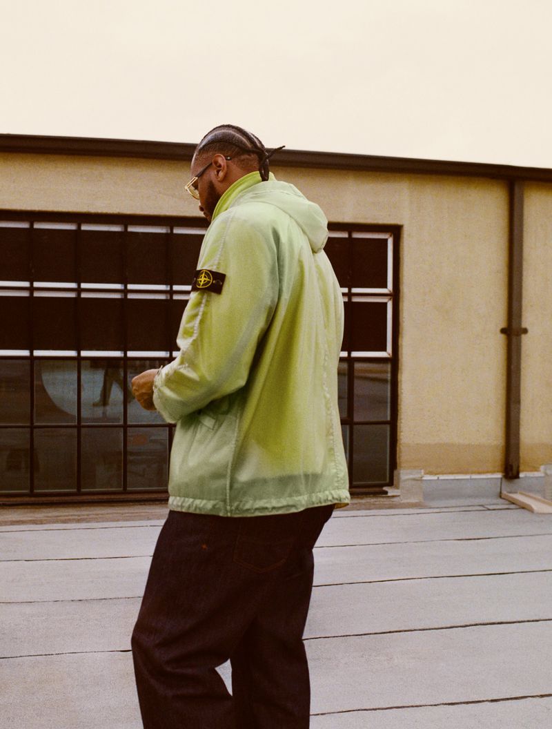 Adult male wearing a lime-green hooded jacket and dark pants is standing outdoors in front of a commercial building on a cloudy day.