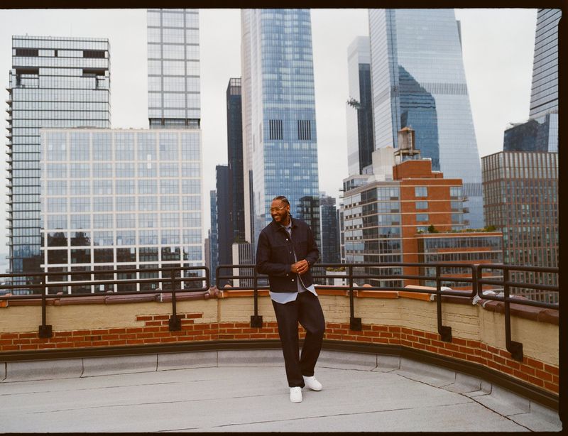Adult man stands on rooftop in city clothes, dark jacket and pants, white sneakers, with a city skyline in the background.