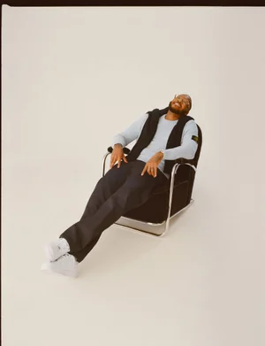 Man laughing while seated in a black armchair against a pale studio backdrop wearing a light sweater and dark trousers