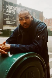 Man wearing tinted glasses and a denim jacket leaning on a green post for Mr Porter editorial