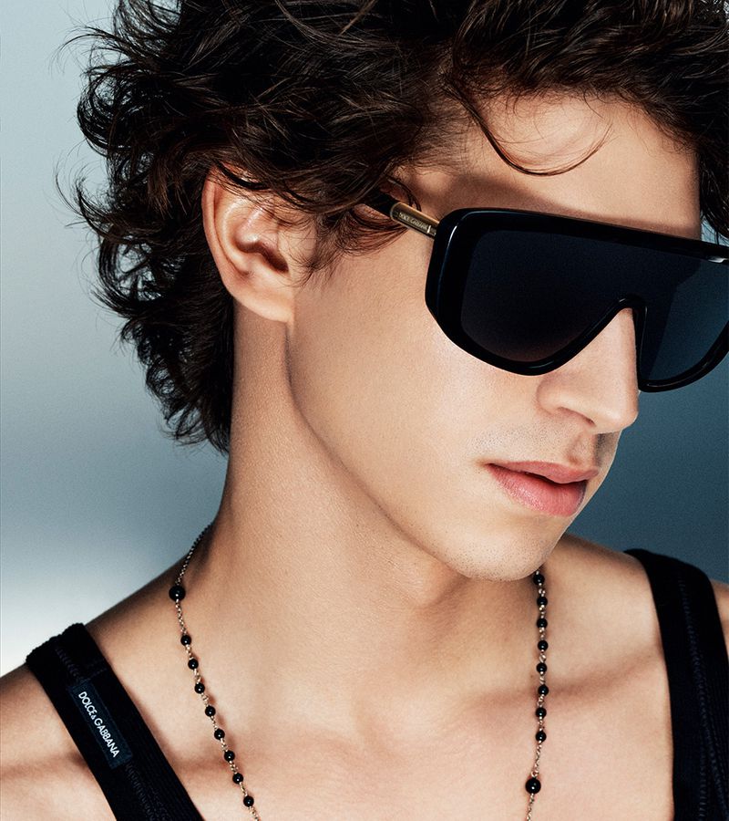 Close-up of a young adult male wearing large black sunglasses and a black tank top, a gold necklace with black beads, and wavy dark hair.