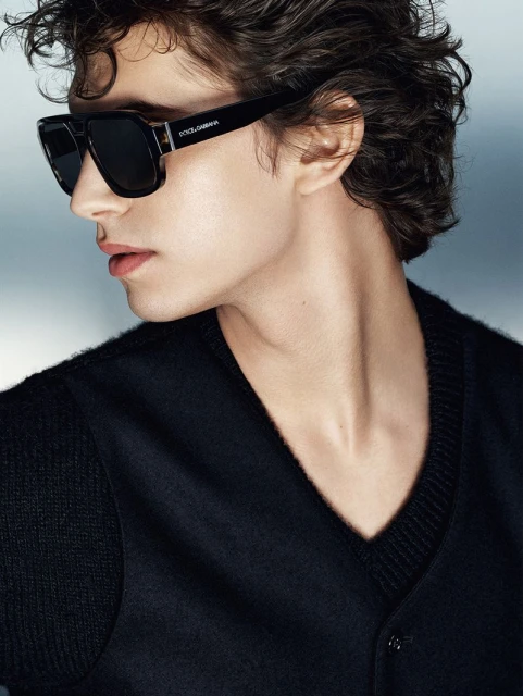 Close-up of a young adult male wearing a black V-neck sweater and Dolce & Gabbana sunglasses against a soft blue gradient background.