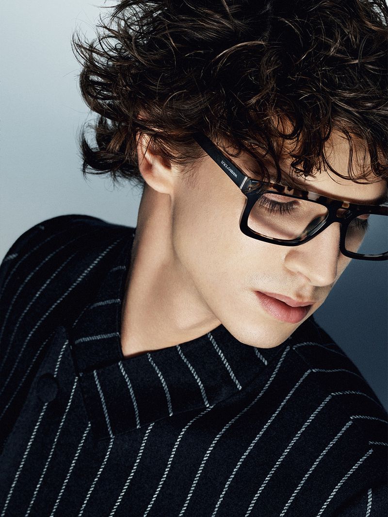 Portrait of a young adult male model with brown curly hair, wearing glasses and a pinstripe blazer, against a gradient light blue background.