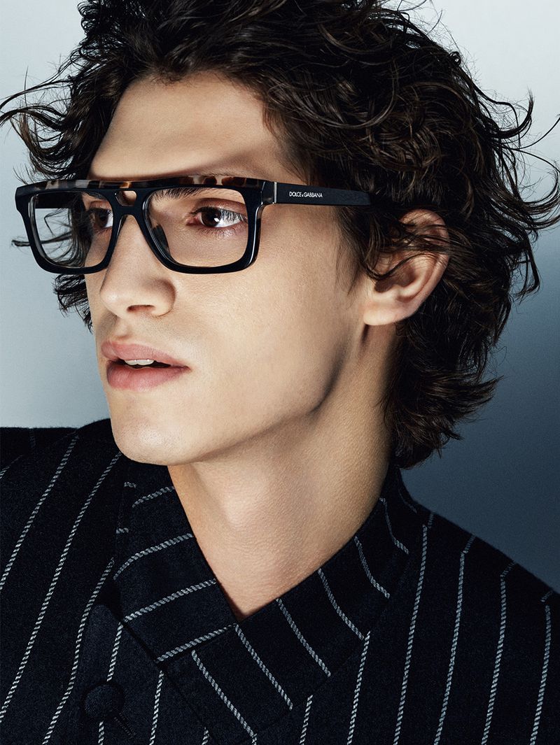 Portrait of an adult man wearing glasses and a striped suit jacket, with a blurred blue gradient background in an indoor setting.