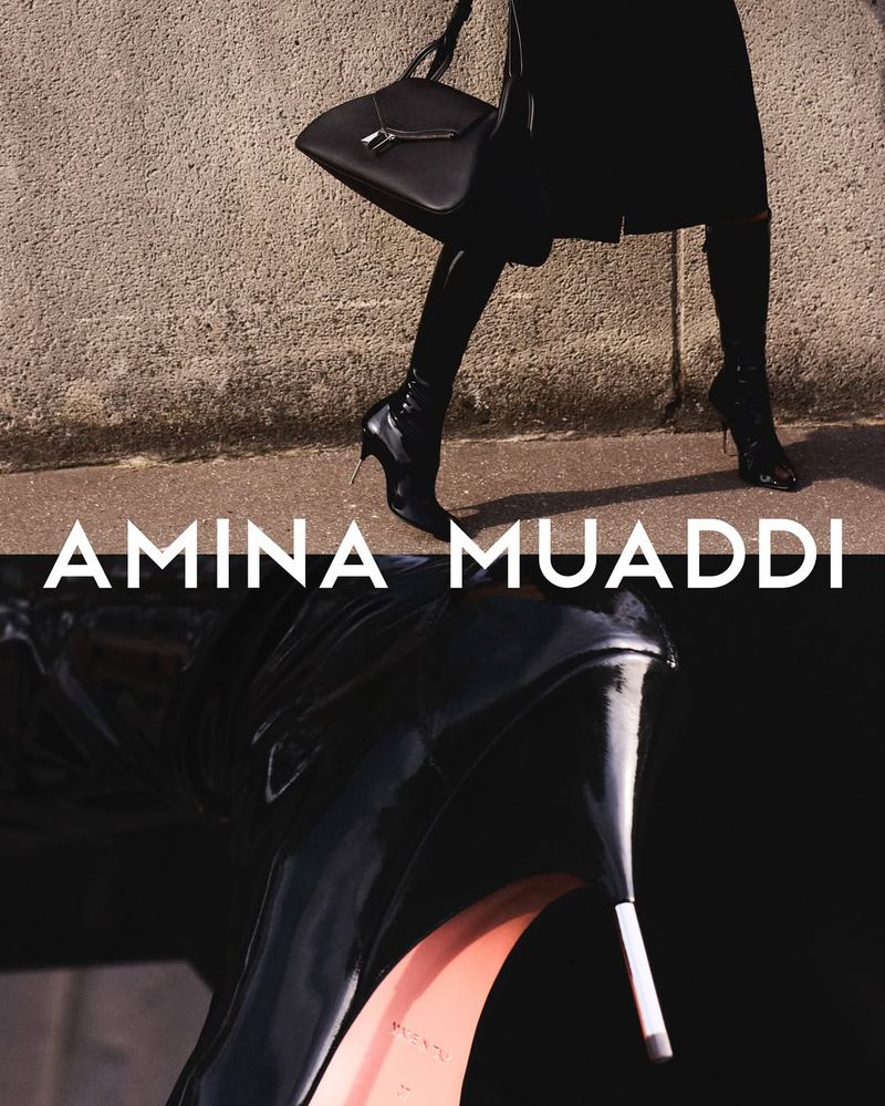 Fashion editorial shot features a woman in black boots and handbag walking by a concrete wall with the brand 'Amina Muaddi' displayed.