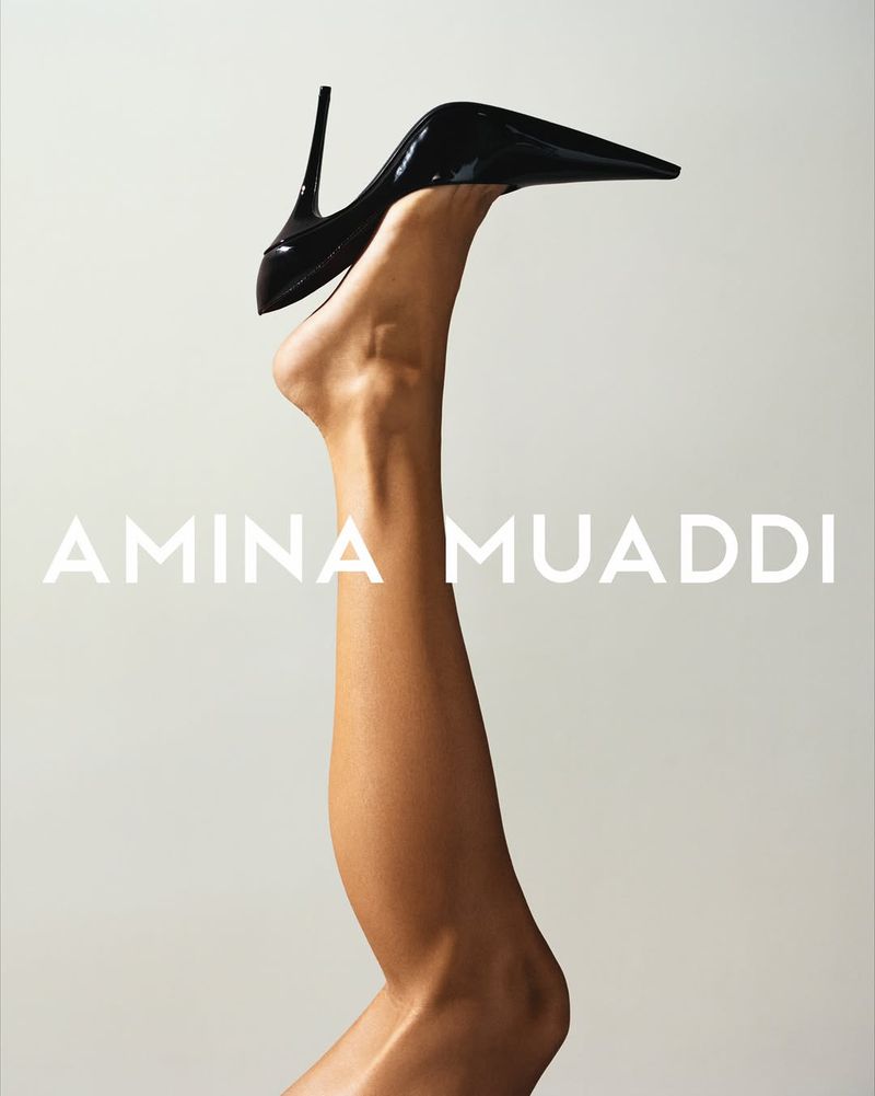 Black high heel shoe on a woman's raised leg against a neutral background, logo AMINA MUADDI visible, advertising fashion and style.