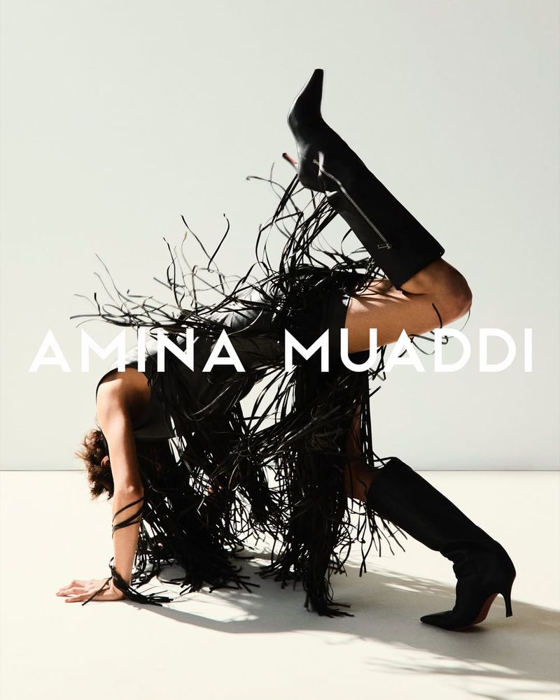 A woman in black fringe outfit and high heel boots is in a handstand position against a white background in a studio setting.