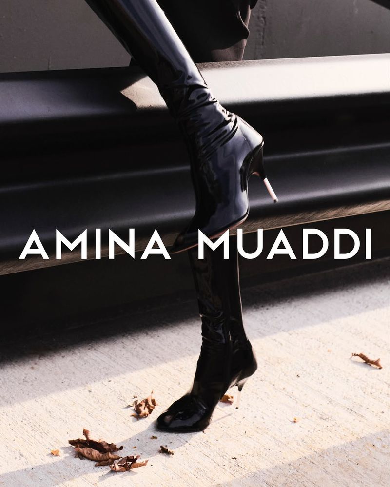 Close-up of black, shiny thigh-high heeled boots with 'Amina Muaddi' text overlay on concrete and dark background, fashion-forward footwear.
