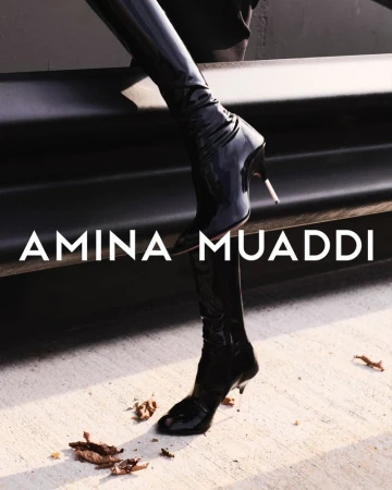 Close-up of black, shiny thigh-high heeled boots with 'Amina Muaddi' text overlay on concrete and dark background, fashion-forward footwear.
