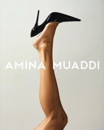 Close-up of a leg raised vertically wearing a glossy black stiletto heel pump against a minimal light grey background.