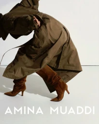 Model in olive trench coat and brown suede heeled boots bending in front of a white seamless background with Amina Muaddi text overlay
