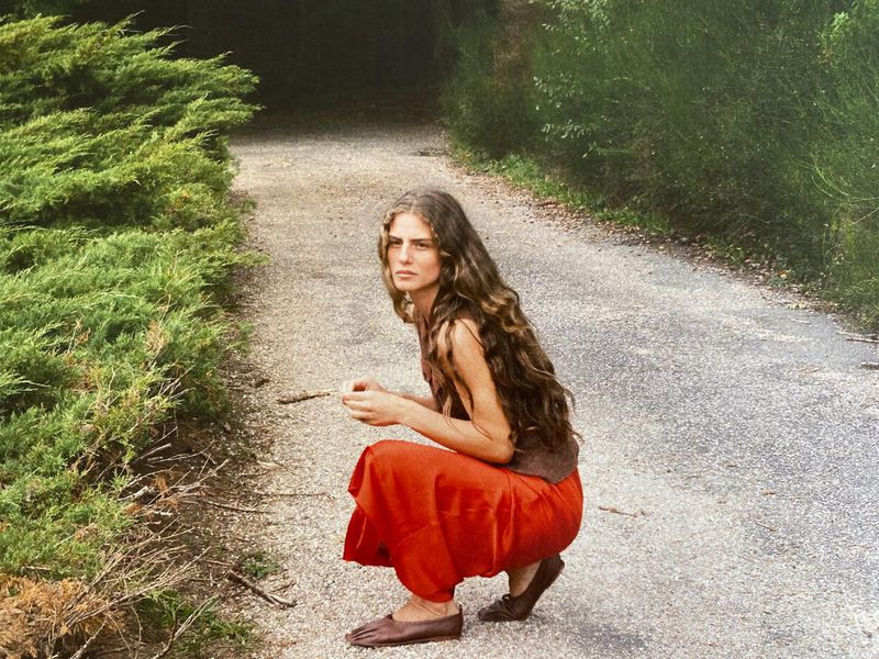 A woman with long brown hair kneels on a path in a rural area. She is wearing an orange skirt and a brown top.