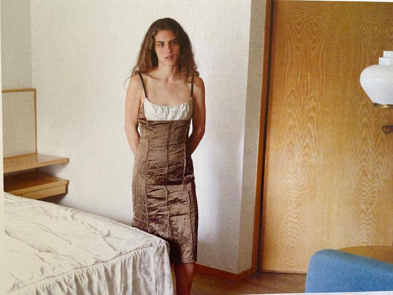 Indoor portrait of an adult woman in a brown dress with a white top, standing next to a bed, with a wooden door in the background.