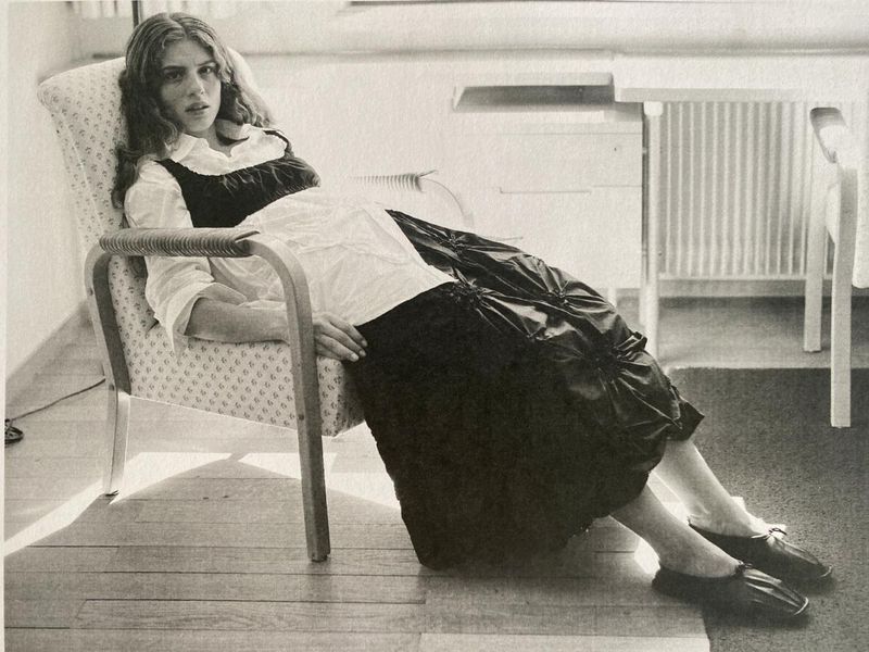 Young woman with wavy hair sits in a patterned chair, wearing a white blouse and black dress, indoors with a wooden floor.