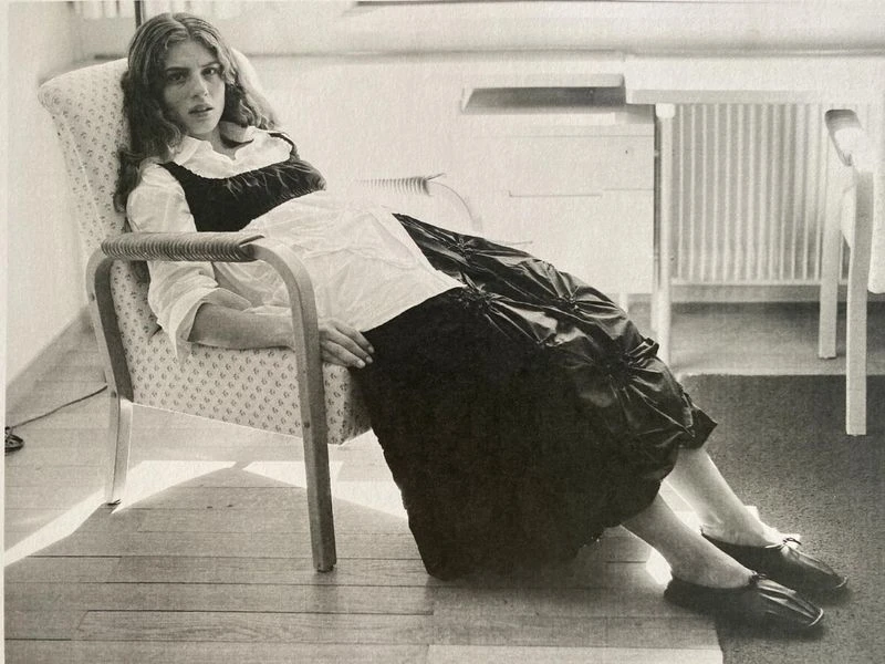 Young woman with wavy hair sits in a patterned chair, wearing a white blouse and black dress, indoors with a wooden floor.