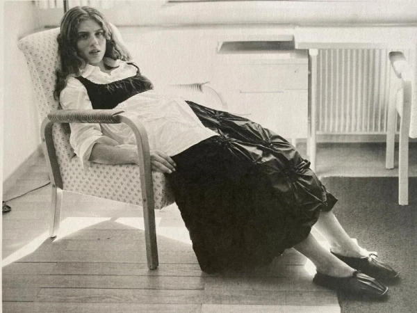 Young woman with wavy hair sits in a patterned chair, wearing a white blouse and black dress, indoors with a wooden floor.