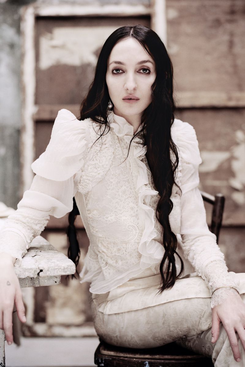 Elegant woman with long dark hair poses in a white lace dress. The soft, muted light highlights textures and vintage tones in the scene.