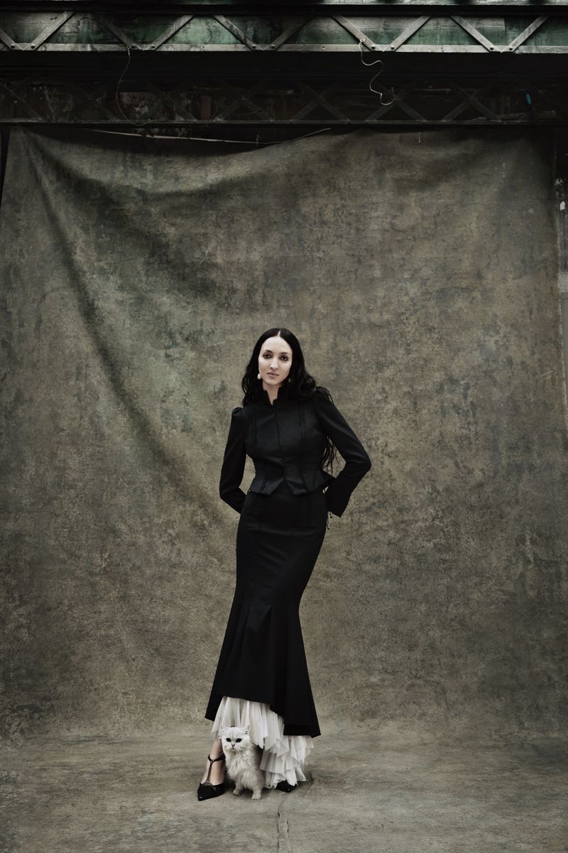 Elegant woman in black formal wear poses with a white cat against a grey studio backdrop, showcasing high fashion and pet photography.