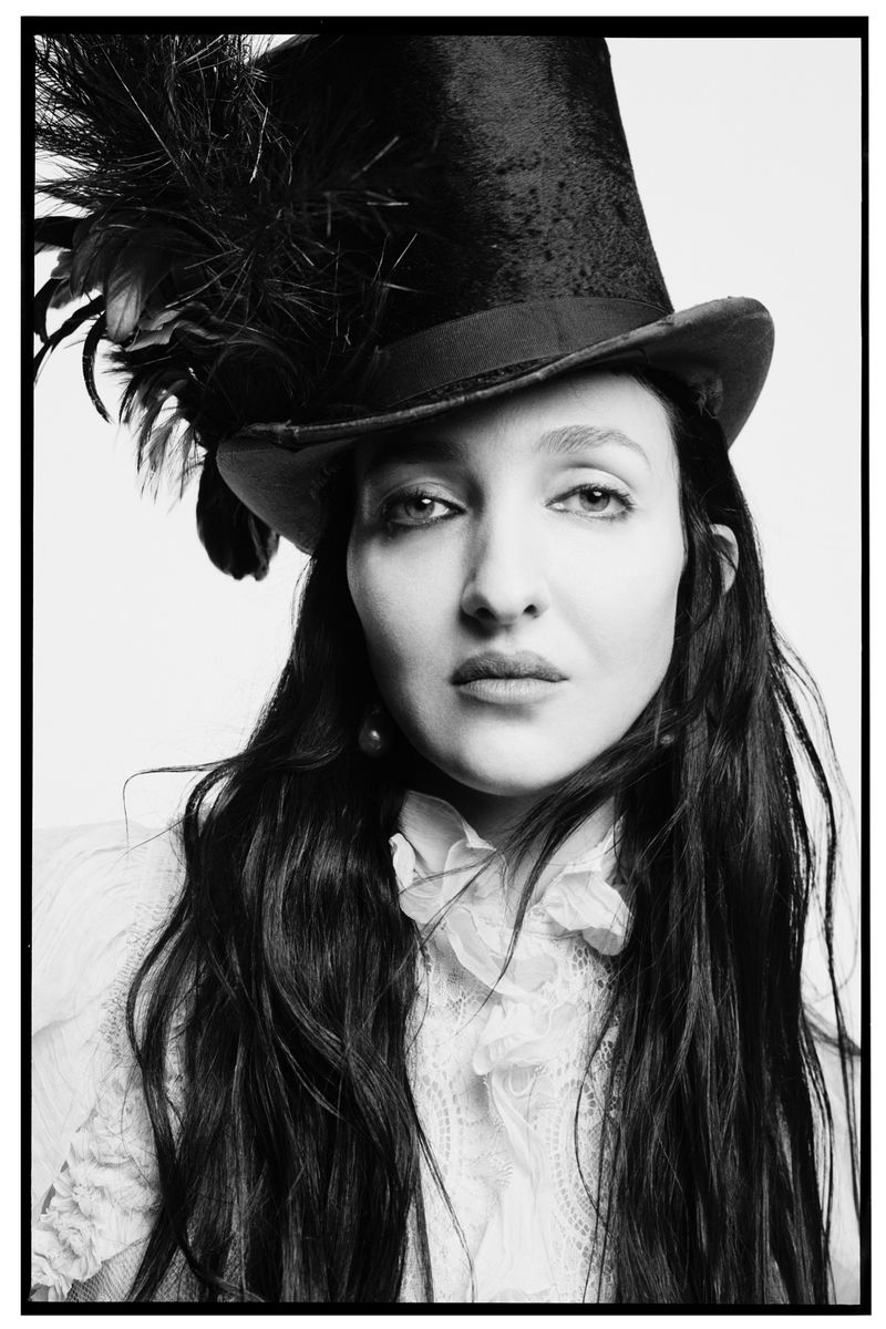 Portrait of a woman with long dark hair wearing a lace blouse and a tall top hat with feather embellishments in a studio setting.
