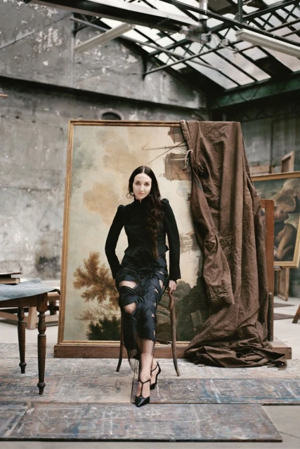 Female model poses in a black outfit in an artist's studio, seated in front of a landscape painting. The studio features old paintings.