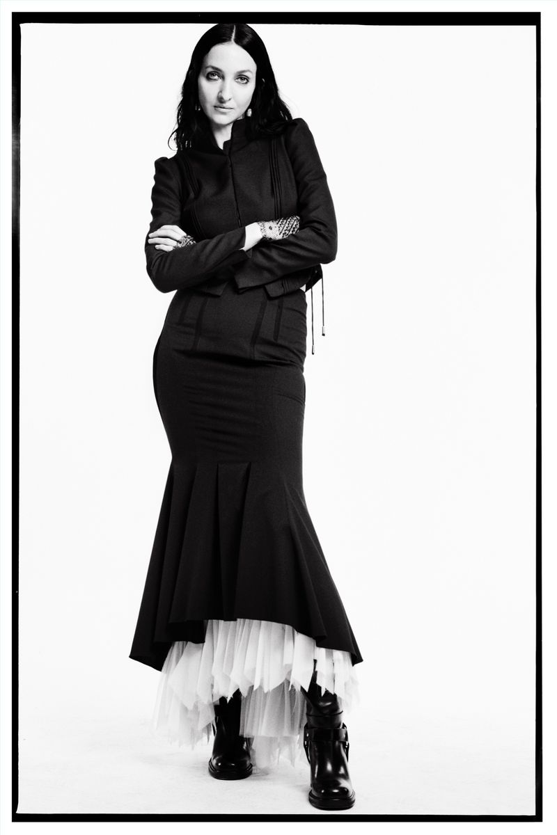 Elegant woman in black jacket and mermaid skirt posing in a studio setting with bright lighting and a white backdrop for a fashion-forward shot.