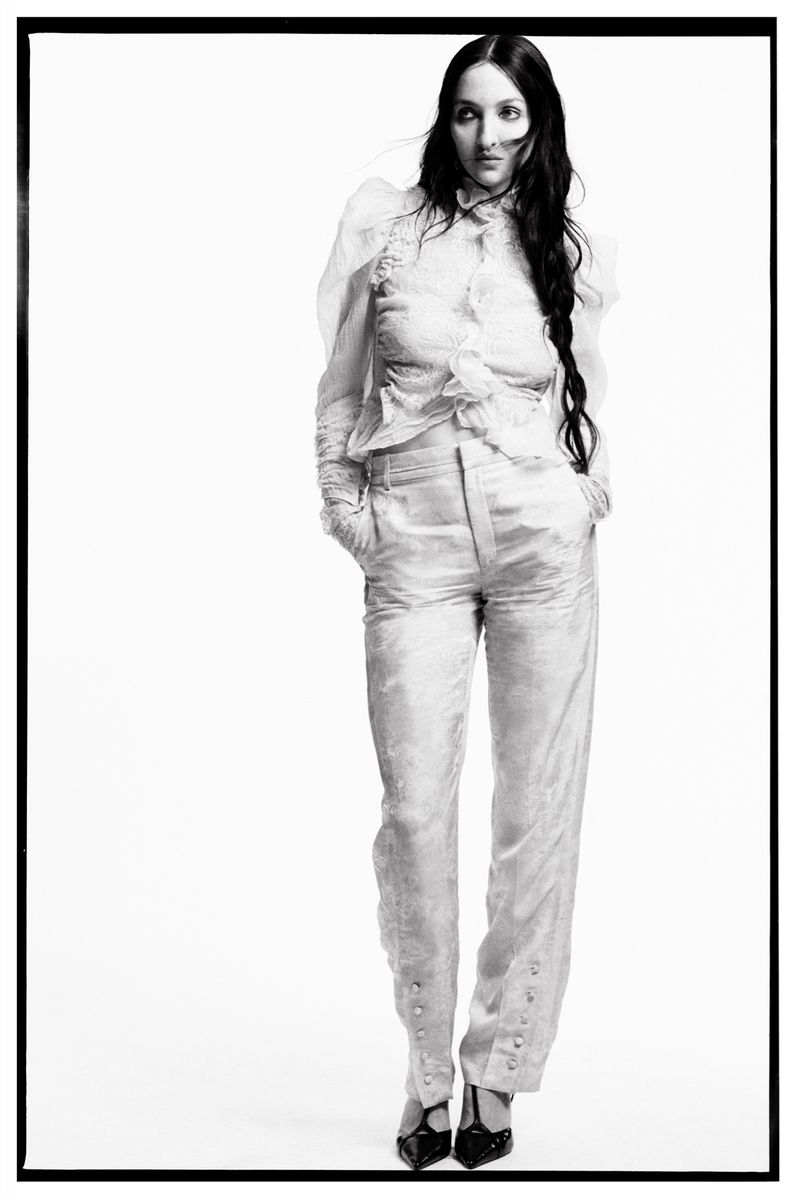 A full shot captures a woman in a light-colored blouse and pants, hands in pockets, posing against a stark white backdrop with a black border.