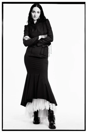 Model Tish Weinstock in black jacket and skirt with white tulle hem standing against a white seamless background