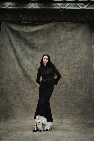 Model Tish Weinstock in a black jacket and long skirt posing with a white cat against a textured backdrop for Zara.