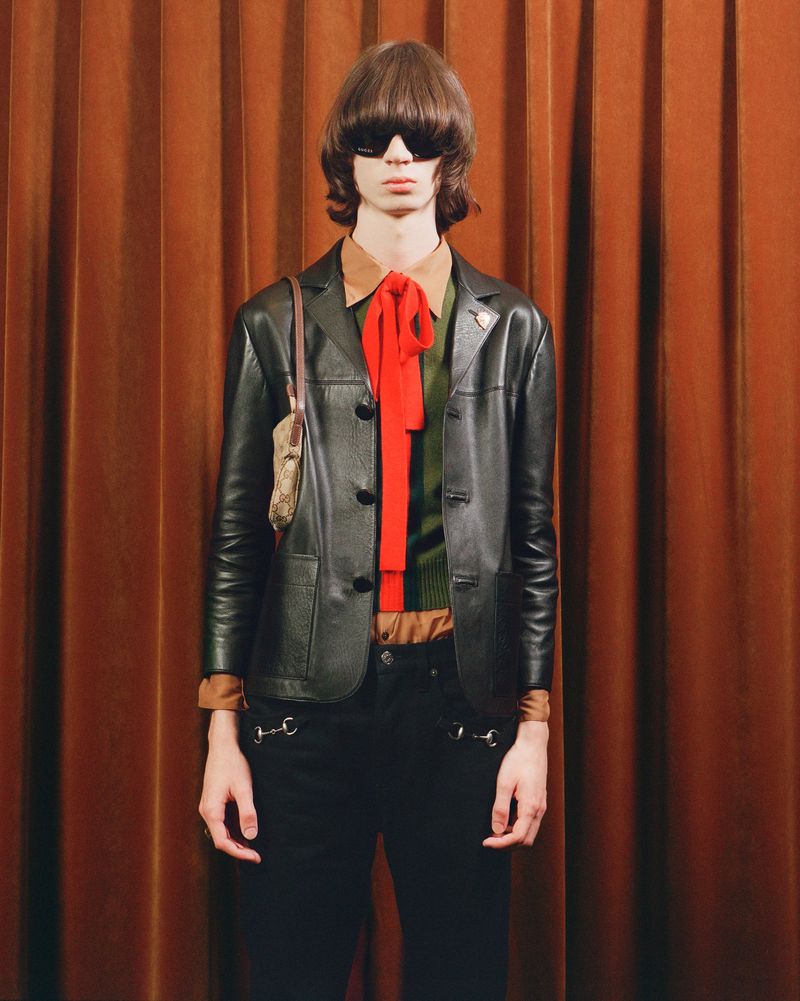 A fair-skinned young adult poses indoors against a dark orange curtain, wearing a black leather jacket, dark pants with silver hardware, and a red scarf.