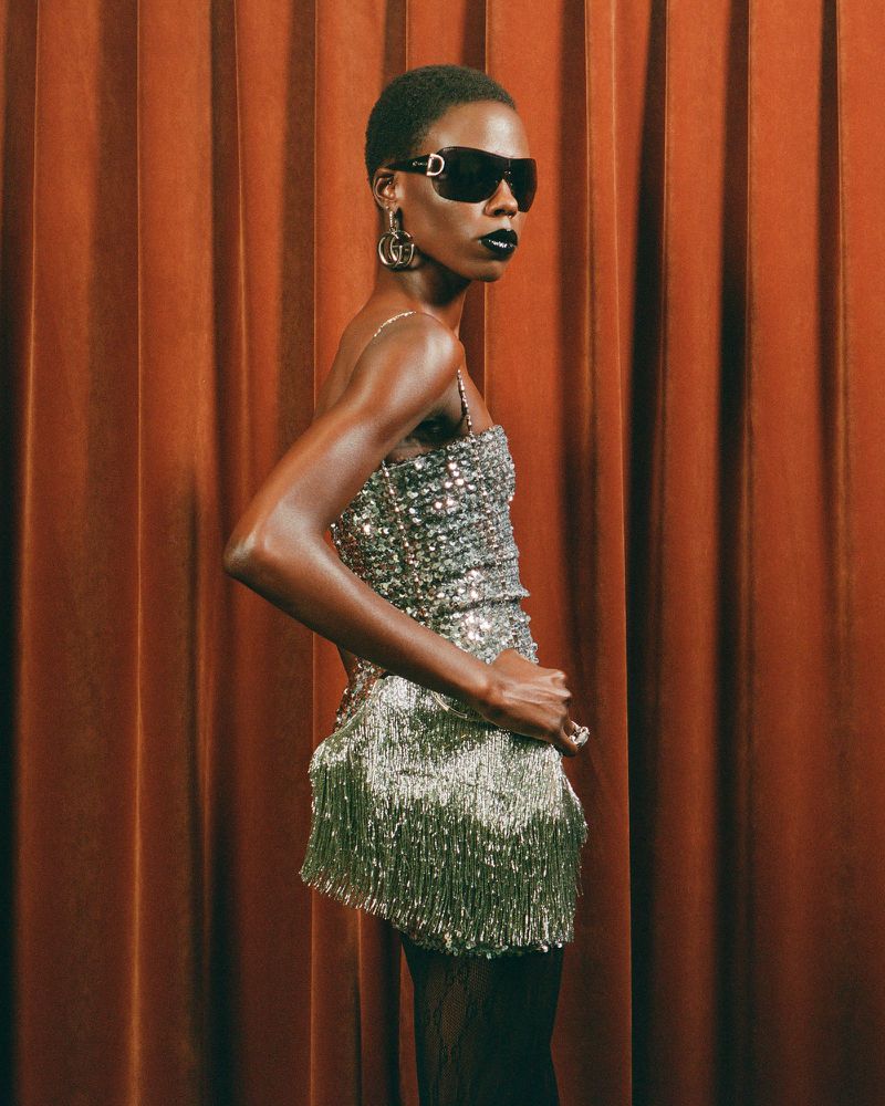 A dark-skinned female model with short hair poses in a shimmering silver fringe dress, sunglasses, and large earrings against a brown curtain backdrop.