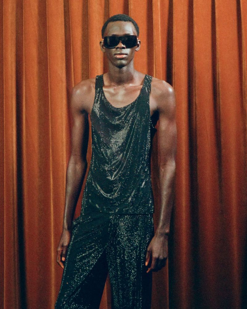 Fashionable Black male model poses in shimmering black tank top and pants against a warm orange curtain backdrop in a studio setting.