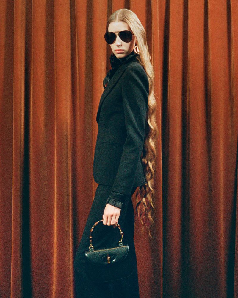 Woman with long blonde hair poses in a dark blazer and skirt with a black purse against a dark orange curtain backdrop indoors.