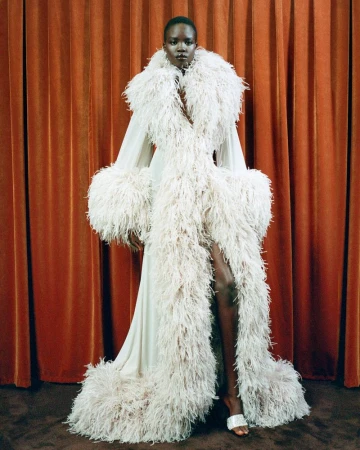 An African American woman in a full-length cream gown trimmed with feathers poses against a copper curtain in a fashion shot.