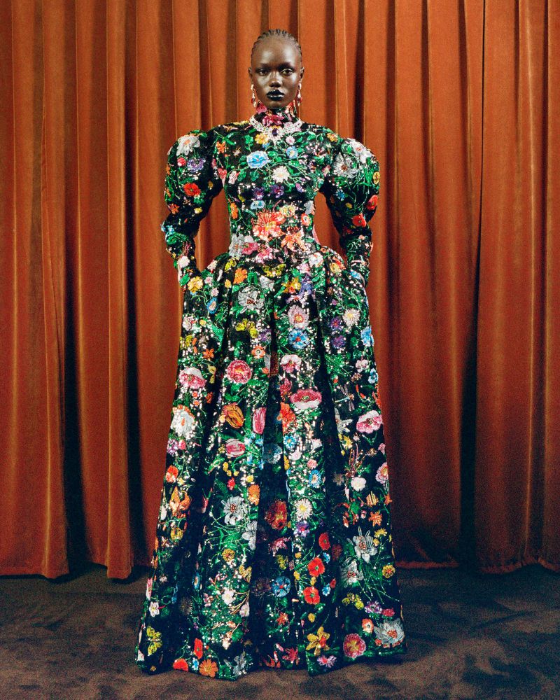 Woman in a long, floral-patterned dress poses in front of an orange curtain; she is wearing gold earrings and has a neutral facial expression.