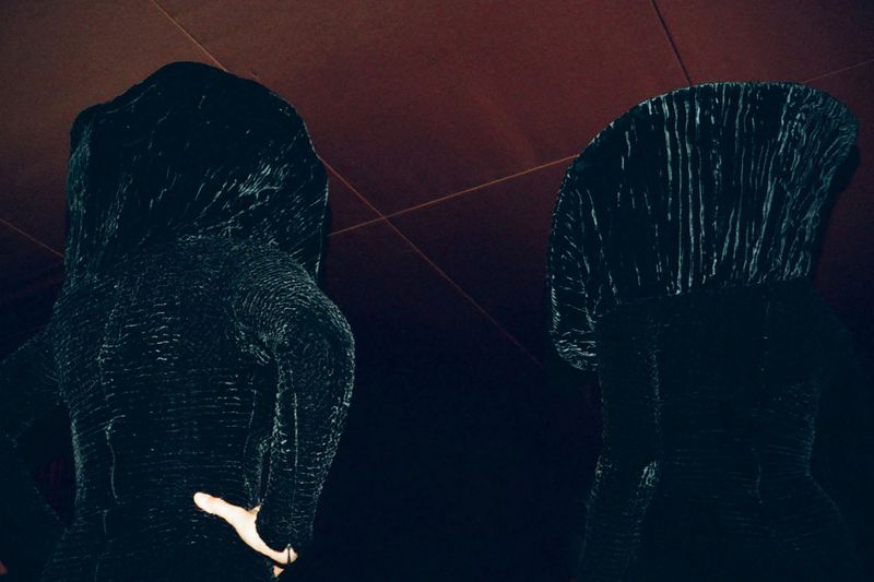 Two women in textured dark hooded outfits stand indoors against a dark background, backs turned in a mysterious, high-fashion style.