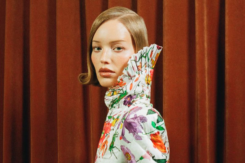 Elegant woman poses in floral top against a rust-colored curtain backdrop, showcasing high fashion in a portrait with neutral tones.