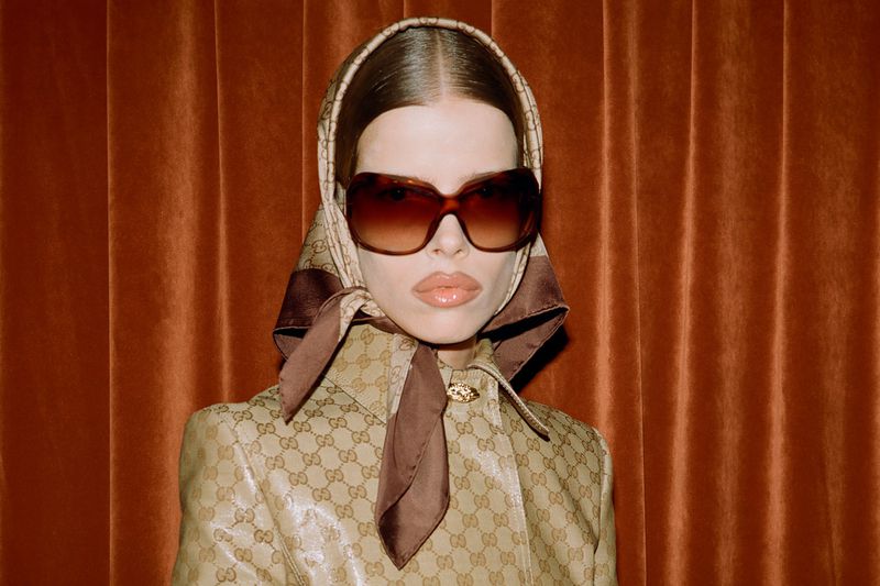 Close-up studio portrait of a woman wearing a patterned headscarf, sunglasses, and jacket, posing against a textured orange backdrop.