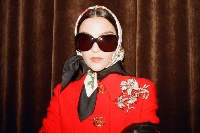 Elegant woman in a red jacket with a floral headscarf and sunglasses poses in front of a draped brown fabric background.