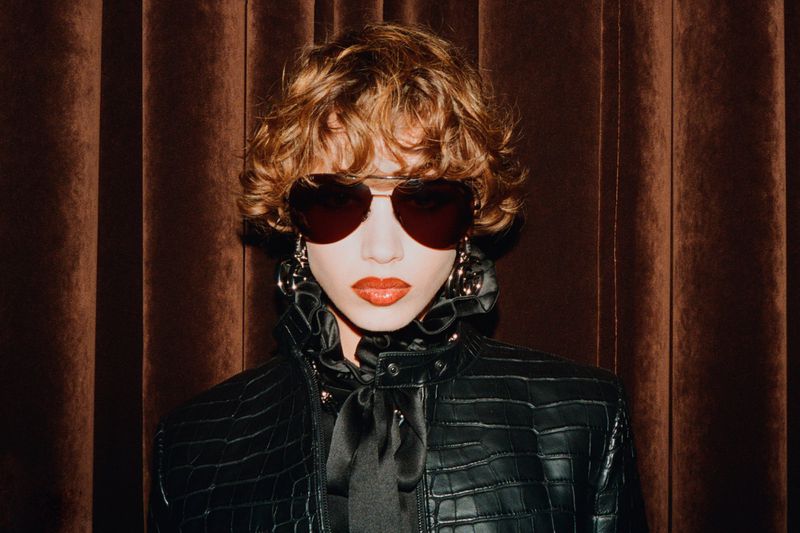 Woman with curly hair, heart-shaped sunglasses, and a black jacket poses against a brown velvet curtain backdrop in soft, diffused lighting.