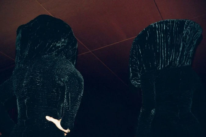 Two figures in dark textured hooded garments viewed from a low angle against a dim ceiling