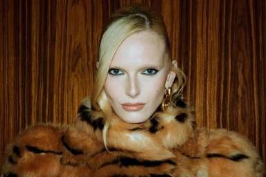 Close-up portrait of a blonde woman wearing a patterned faux fur coat against a dark wood grain background.