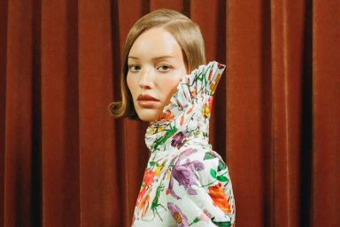 Model wearing a white floral high-neck garment posing against a vertical brown velvet curtain background