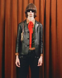 Model wearing a black leather jacket, red necktie, and dark sunglasses against a pleated orange curtain background.