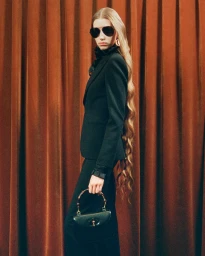 Model with very long wavy hair wearing dark sunglasses, black blazer, and holding a handbag against a rust-colored curtain background.